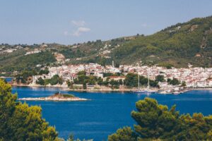 A wide shot of white and brown houses near blue ocean surrounded by mountains with trees in Skiathos, Greece